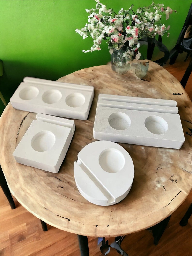 May include: Four white ceramic coasters with different shapes and sizes. The coasters are on a wooden table with a vase of white flowers in the background.
