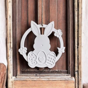 Könnte beinhalten: Ein weißer Holz-Osterhasen-Kranz mit einem Herzen und Eiern. Der Kranz hängt an einer Holztür.