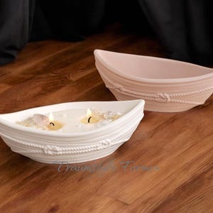 Könnte beinhalten: Zwei weiße Kerzenhalter aus Keramik in Form von Booten mit Seildetails. Die Boote sind mit weißem Wachs gefüllt und haben drei kleine Teelichter, die darin brennen. Die Boote stehen auf einer Holzoberfläche.