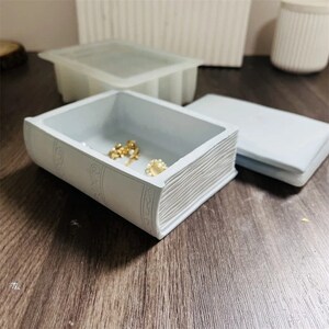 Könnte beinhalten: Eine weiße, buchförmige Schmuckschatulle mit einem detaillierten Rücken-Design. Die Schachtel ist geöffnet und enthält ein paar Schmuckstücke. Die Schachtel steht auf einer Holzoberfläche.