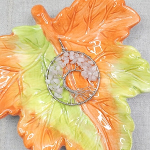 Op de afbeelding: Een zilveren draad hanger in de vorm van een boom, gevuld met kleine, lichtroze en witte stenen. De hanger ligt op een keramische blad-vormige schaal met oranje, gele en groene tinten.