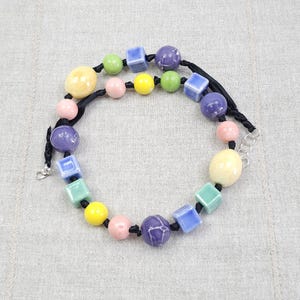Op de afbeelding: Een ketting met een zwart koord geregen met diverse keramische kralen in verschillende vormen en kleuren, waaronder ronde, ovale en kubusvormen in tinten geel, roze, groen, blauw en paars, afgewerkt met een zilverkleurige metalen sluiting.