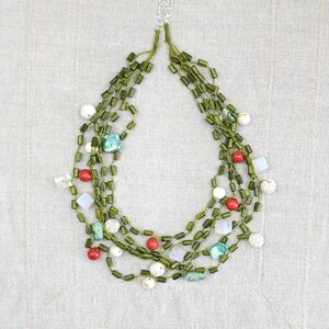 Op de afbeelding: Een meerlagige ketting met groene, rode, witte en turquoise kralen. De ketting heeft een zilveren sluiting en bestaat uit kralen in verschillende vormen en maten, waardoor een gelaagd effect ontstaat. De kralen zijn geregen op een groen koord.