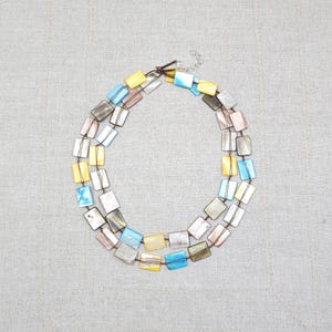 Puede incluir: Un collar de dos hilos con cuentas rectangulares e iridiscentes en tonos amarillos, azules, rosas y grises. Las cuentas están ensartadas en un cordón marrón y aseguradas con un cierre plateado. El collar se exhibe sobre un fondo neutro.