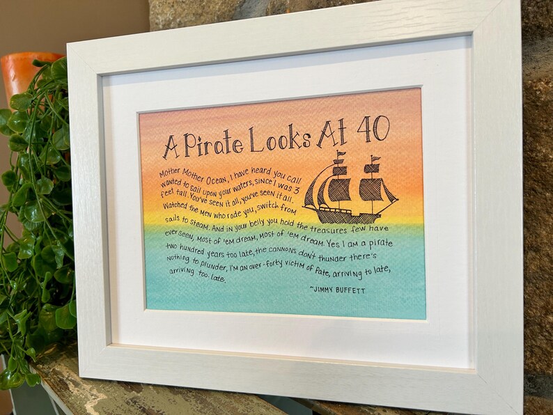 May include: A framed print with a watercolor background in shades of yellow, orange, green, and blue. The print features a black and white illustration of a pirate ship with the text "A Pirate Looks At 40" and a quote from Jimmy Buffett: "Mother Mother Ocean, I have heard you call wanted to sail upon your waters, since I was 3 Feel tall. You've seen it all, you've seen it all. Watched the men who rade you, switch from sails to steam. And in your belly you hold the treasures few have ever seen. Most of 'em dream, Most of 'em dream. Yes I am a pirate two hundred years too late, the cannons don't thunder there's nothing to plunder, I'm an over-forty victim of fate, arriving to late, arriving too late."