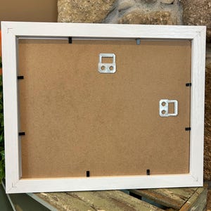 May include: The back of a white picture frame with a brown cardboard backing. Two metal hanging hooks are attached to the back of the frame.
