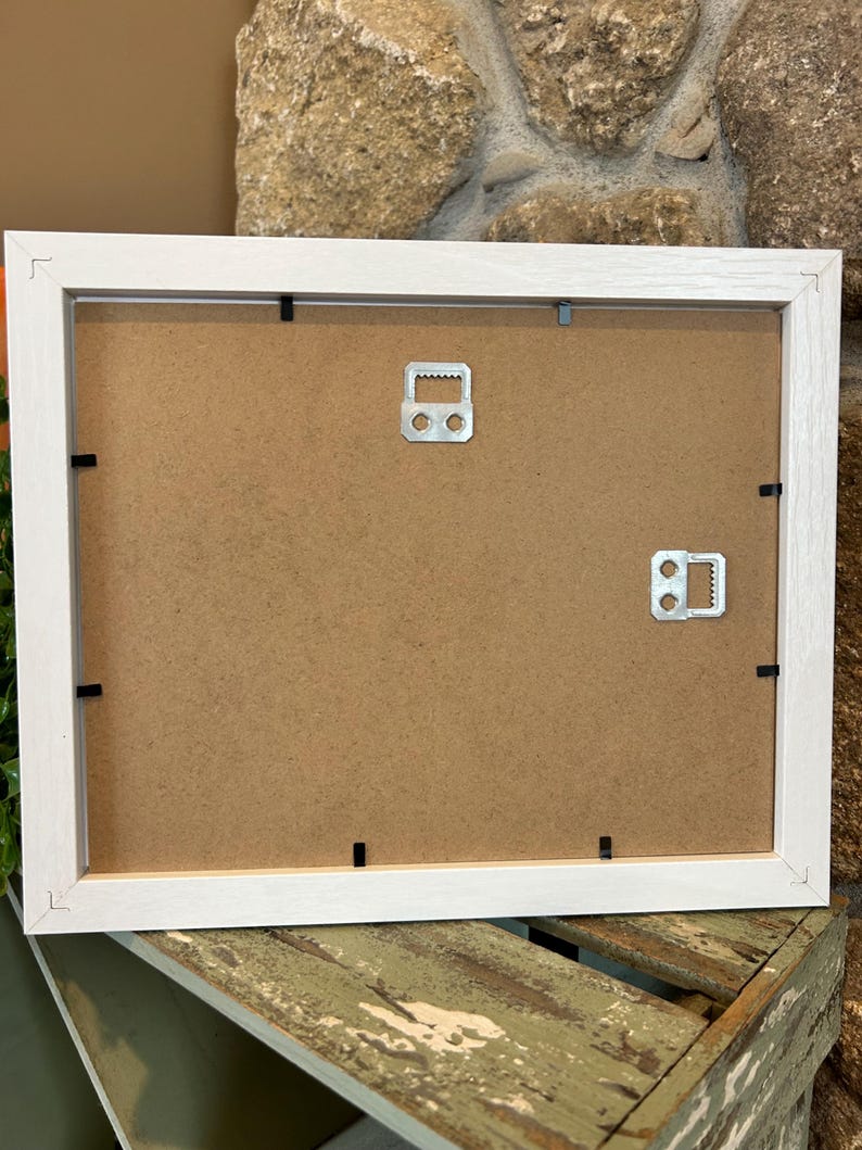 May include: The back of a white picture frame with a brown cardboard backing. Two metal hanging hooks are attached to the back of the frame.