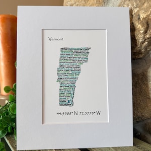 May include: A framed print featuring the outline of Vermont, filled with the names of towns and landmarks in a green font. The word "Vermont" is at the top, with coordinates 44.5588° N 72.5778° W at the bottom. The print is in a white frame.
