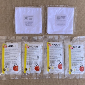 Op de afbeelding: Vier pakjes witte Nigari-vlokken, elk 100 gram, met een geel etiket met de tekst "Nigari" en "100g". Er zijn ook drie witte mousseline kaasdoeken afgebeeld, elk met een geel etiket met de tekst "Mussola Cheese Cloth".