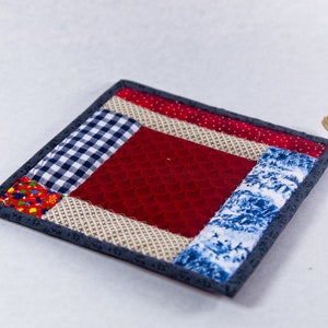 Puede incluir: Un posavasos cuadrado acolchado con un diseño de patchwork rojo, blanco y azul. El posavasos tiene un borde gris y un lazo para colgar.