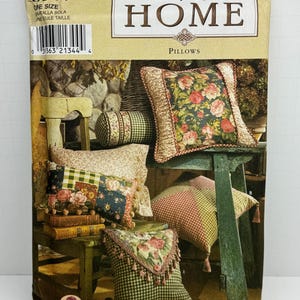 Puede incluir: Patrón de costura Simplicity Home número 8044 para almohadas. La portada del patrón presenta una foto de cinco almohadas en varios colores y patrones, incluyendo diseños florales, a cuadros y de patchwork. Las almohadas están dispuestas en una silla y una mesa de madera.