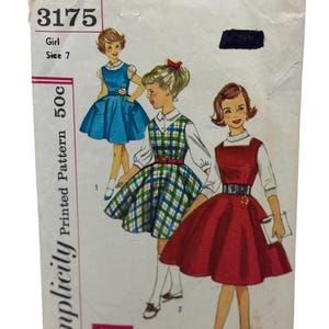 Op de afbeelding: Vintage Simplicity naaipatroon voor een meisjesjurk. Het patroon is nummer 3175 en is voor maat 7. Het patroon toont drie verschillende stijlen jurken, een blauwe, een geruite en een rode.