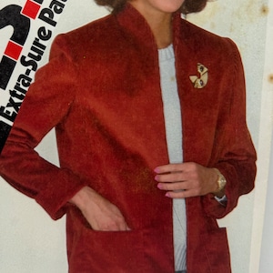May include: A rust-colored corduroy jacket with a mandarin collar and a zipper closure. The jacket has a pocket on the left side and a decorative pin on the right side.