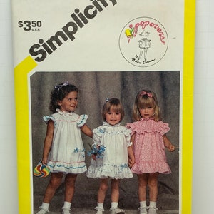 Puede incluir: Patrón de costura Simplicity número 6352 para vestidos de niñas. El patrón incluye tres estilos diferentes de vestidos, todos con cuellos y mangas con volantes. Los vestidos se muestran en tres niñas pequeñas, una con un vestido blanco con ribetes azules, otra con un vestido blanco con ribetes de encaje y otra con un vestido rosa con estampado floral.