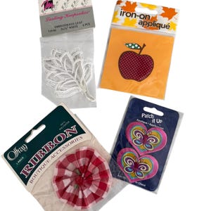 May include: Assortment of craft supplies including an embroidered white leaf, an iron-on apple appliqué, a red and white gingham ribbon, and two heart-shaped butterfly patches. The packaging includes the text "Lasting Keepsakes", "Iron-on appliqué", "Ofray Ribbon", and "Patch it Up".