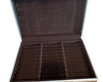 May include: A brown rectangular carrying case with a silver metal frame and a brown handle. The case is open, revealing a dark brown interior lined with velvet-like material, featuring three vertical columns of molded slots for storage.
