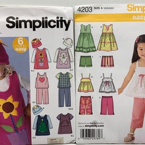 Puede incluir: Dos patrones de costura para ropa de niños. El patrón de la izquierda es para un vestido y un sombrero con temática floral. El patrón de la derecha es para una blusa blanca con un lazo rosa y pantalones rosas. Ambos patrones son de Simplicity y son fáciles de coser.
