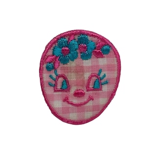 May include: Pink and white gingham fabric with a smiling face embroidered in pink thread. The face has blue eyes and a flower crown with blue flowers.