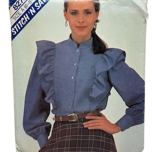 May include: A vintage sewing pattern cover featuring a woman wearing a blue denim blouse with ruffled sleeves and a brown plaid skirt. The blouse has a button-down front and a high collar. The text "STITCH 'N SAVE" is visible.