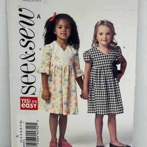 May include: A sewing pattern for girls' dresses. The pattern features two dress styles: one with a floral print and lace trim, and the other in a black and white houndstooth pattern. The pattern includes sizes for children aged 2 to 8 years old.