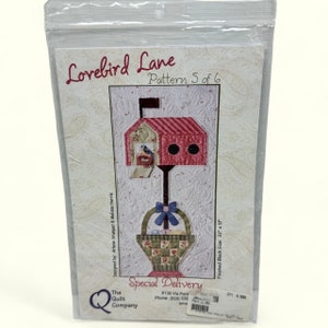 May include: A quilting pattern for a mailbox with a birdhouse on top and a basket underneath. The pattern is called "Lovebird Lane" and is pattern 5 of 6. The finished block size is 56 cm by 28 cm.