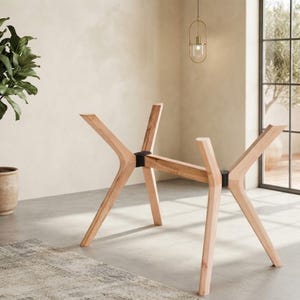 May include: A light-colored wooden table base featuring four angled legs that extend outward from a central horizontal beam. The structure is connected by two black metal joints that secure the wooden components together in a stable frame.