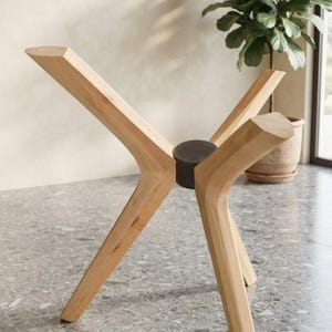 May include: A table base featuring four light-toned wooden legs arranged in an X-shape, connected by a central dark gray cylindrical metal hub. The legs extend outward at an angle, providing a stable foundation for a tabletop surface.