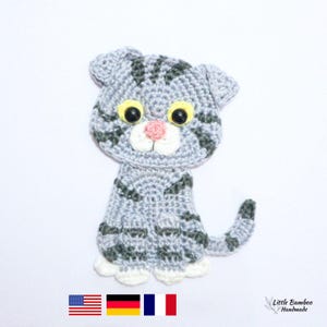 May include: Crocheted gray cat with dark gray stripes, yellow eyes, and a pink nose. The handmade cat has white paws and a curled tail. The cat is sitting upright. The image includes the text "Little Bamboo Handmade".