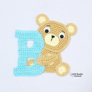 Puede incluir: Un oso de peluche tejido a ganchillo en tonos beige y crema, sosteniendo una letra "B" azul claro. El oso tiene ojos negros y una nariz negra. La imagen est&aacute; sobre un fondo blanco. El texto "Little Bamboo Handmade" es visible.