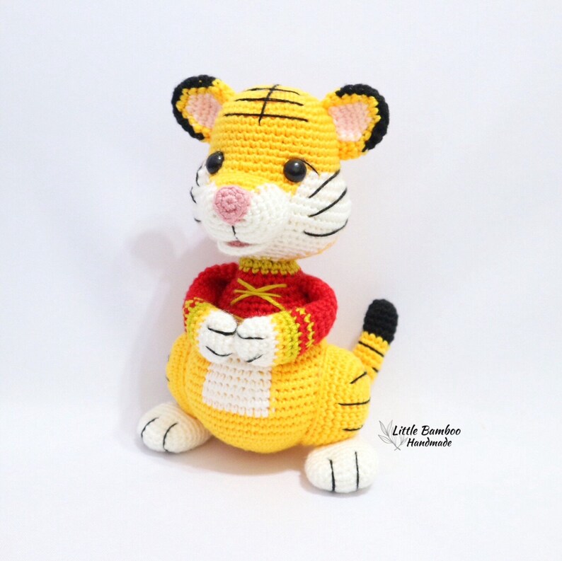 May include: A handmade crochet tiger doll in yellow, white, black, and red. The tiger has a yellow body, white face, and black stripes. It wears a red shirt with yellow accents and has a black and yellow striped tail.