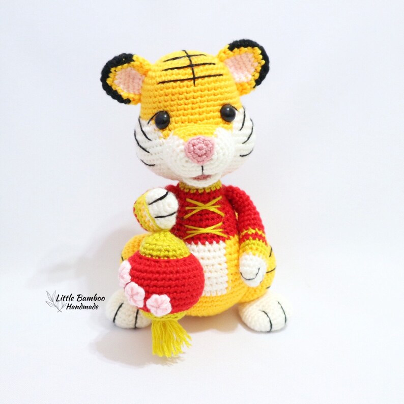 May include: A handmade crochet tiger doll in yellow, white, red, and black. The tiger holds a red and yellow lantern with pink flower accents. The doll has black eyes and a pink nose. The text "Little Bamboo Handmade" is in the lower left corner.