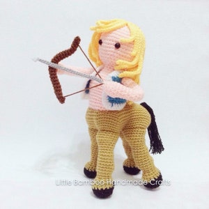 Pode incluir: Um brinquedo de pelúcia de crochê de um centauro com um arco e flecha. O centauro tem um torso humano com cabelo loiro e um corpo de cavalo. O brinquedo está vestindo um colete azul e rosa. O brinquedo está em pé sobre uma superfície branca. "Little Bamboo Handmade Crafts" está escrito na superfície.