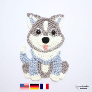 以下が含まれることがあります： グレー、白、青の糸を使った手作りのかぎ針編みのハスキー犬。犬は黒い目とピンクの舌をしています。画像には、アメリカ、ドイツ、フランスの国旗が含まれています。犬は座っています。