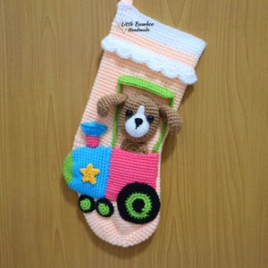 May include: A crocheted Christmas stocking featuring a pink, blue, green and yellow train design. A brown crocheted dog is sitting in the train. The stocking has a white crocheted cuff with a scalloped edge. The text "Little Bamboo Handmade" is on the cuff.