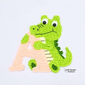 Puede incluir: Un parche de ganchillo hecho a mano con un cocodrilo verde que sostiene la letra "A" de color melocot&oacute;n. El cocodrilo tiene ojos negros y una boca negra. El parche est&aacute; sobre un fondo blanco. El texto "Little Bamboo Handmade" est&aacute; en la esquina inferior derecha.