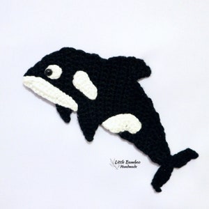 May include: Black and white crocheted orca whale with a black eye. The orca is in a jumping pose. The orca is handmade and has the text "Little Bamboo Handmade" at the bottom.