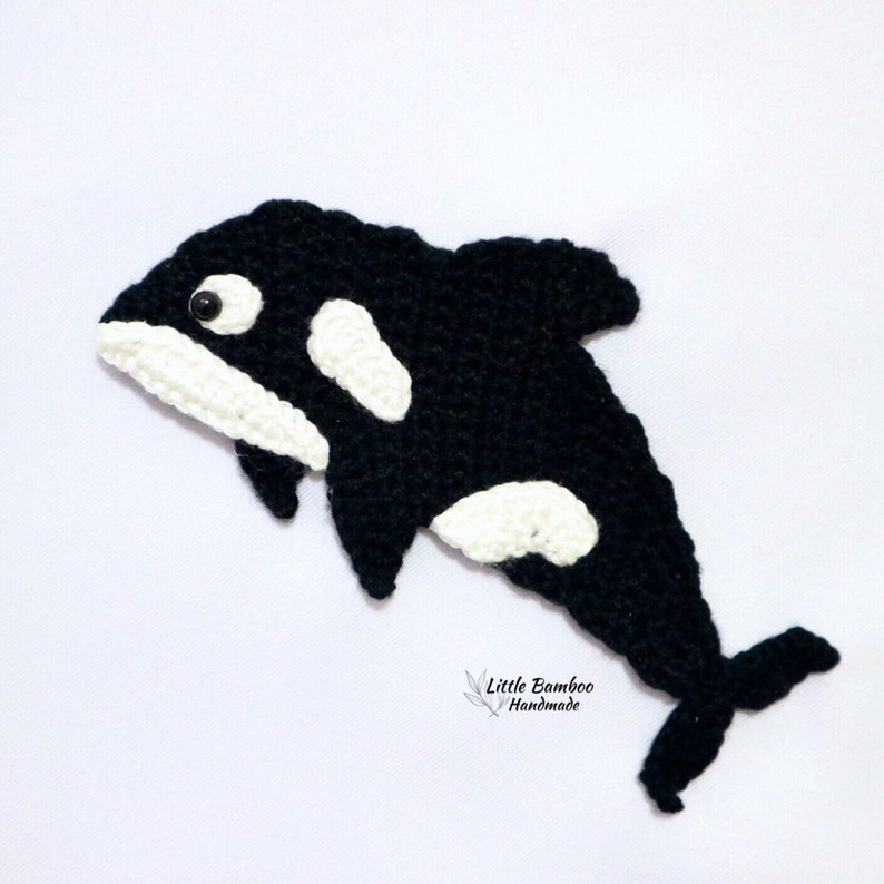 May include: Black and white crocheted orca whale with a black eye. The orca is in a jumping pose. The orca is handmade and has the text "Little Bamboo Handmade" at the bottom.