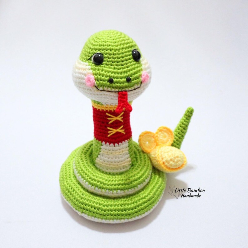 May include: A green crocheted snake with a red and yellow collar and a yellow flower on its tail. The snake has black eyes and a pink nose. The snake is sitting on a white surface. "Little Bamboo Handmade" is written in the bottom right corner of the image.