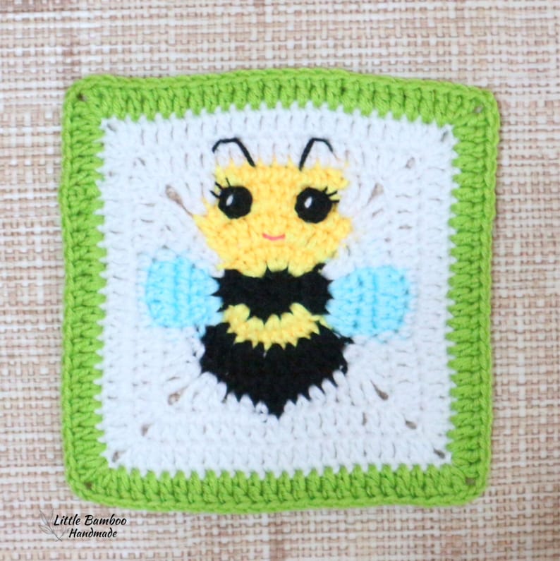May include: A crocheted square featuring a bee with black and yellow stripes, blue wings, and a white face. The square has a green border.