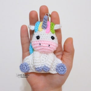 Pode incluir: Um chaveiro de unicórnio branco de crochê com uma crina e um chifre arco-íris. O unicórnio tem bochechas cor-de-rosa, olhos pretos e pés roxos.