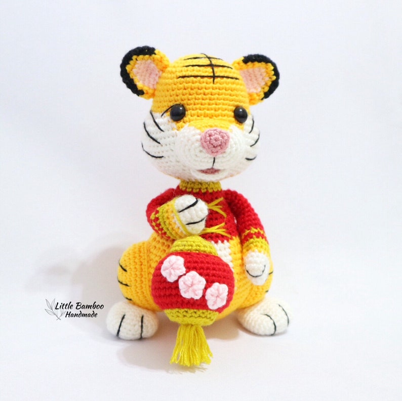 May include: A handmade crochet tiger doll in yellow, white, black, and red. The tiger holds a red lantern with pink flower accents and a yellow tassel. The doll has black eyes, a pink nose, and black stripes.