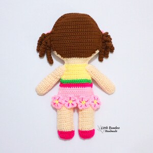 May include: A crocheted doll with brown hair, a yellow and green top, a pink skirt with pink flowers, and pink shoes. The doll is handmade and has a tag that says "Little Bamboo Handmade".