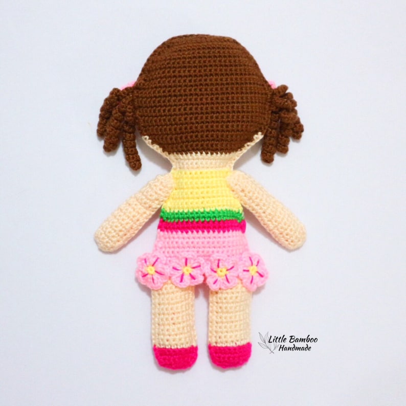 May include: A crocheted doll with brown hair, a yellow and green top, a pink skirt with pink flowers, and pink shoes. The doll is handmade and has a tag that says "Little Bamboo Handmade".