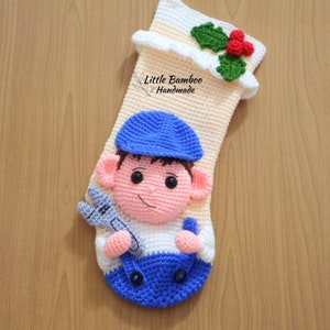 May include: A crocheted Christmas stocking featuring a cartoon character wearing a blue hat and overalls, holding a wrench. The stocking is decorated with a green holly sprig and red berries. The text "Little Bamboo Handmade" is visible on the stocking.