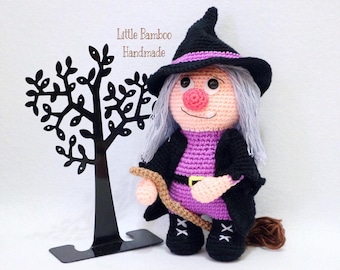 May include: A crocheted witch doll wearing a black hat and purple dress, holding a broom and a stick. The doll is standing on a black metal tree stand. The text "Little Bamboo Handmade" is visible in the image.