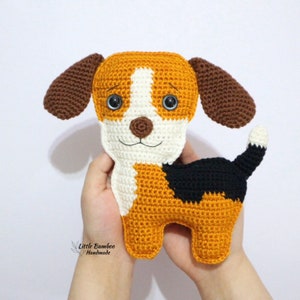 May include: Crocheted beagle dog plush toy with brown ears, white and orange fur, and black markings. The toy has large blue eyes and a brown nose.