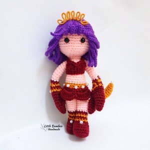 May include: Crocheted doll with purple hair, a red and gold outfit, and a gold crown. The doll has a pink face and brown eyes.