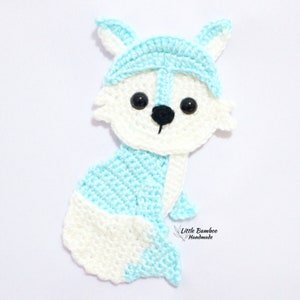 May include: A crocheted blue and white fox applique with black eyes and a black nose. The fox is sitting with its tail curled around its body. The text "Little Bamboo Handmade" is visible in the bottom right corner of the image.