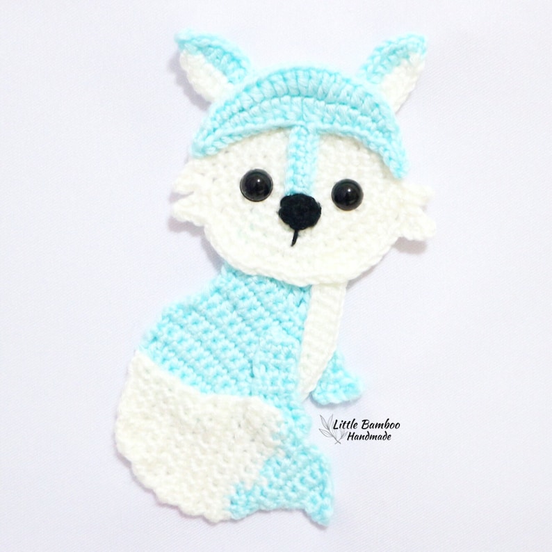 May include: A crocheted blue and white fox applique with black eyes and a black nose. The fox is sitting with its tail curled around its body. The text "Little Bamboo Handmade" is visible in the bottom right corner of the image.