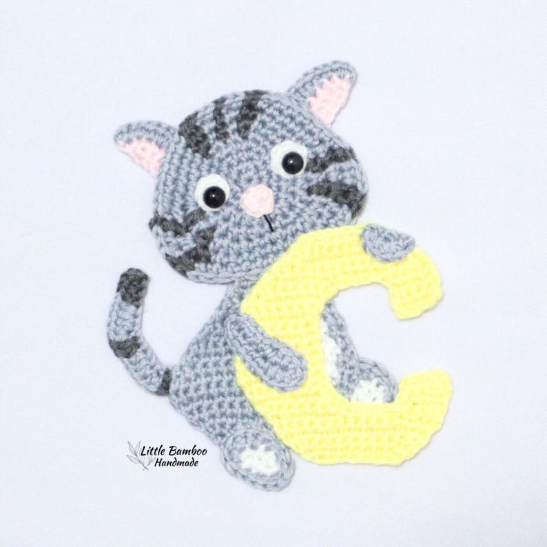 Puede incluir: Un gato de ganchillo gris con rayas grises oscuras, sosteniendo la letra "C" amarilla. El gato tiene orejas internas rosas y una nariz rosa. El texto "Little Bamboo Handmade" est&aacute; en la esquina inferior izquierda.